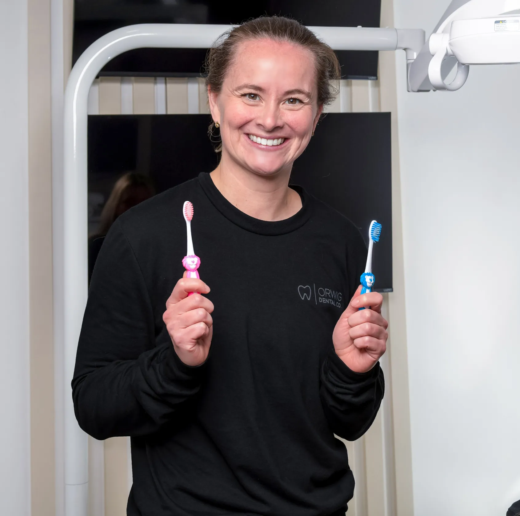 Smiling woman in black shirt holding a pink and a blue children's toothbrush in a dental office.