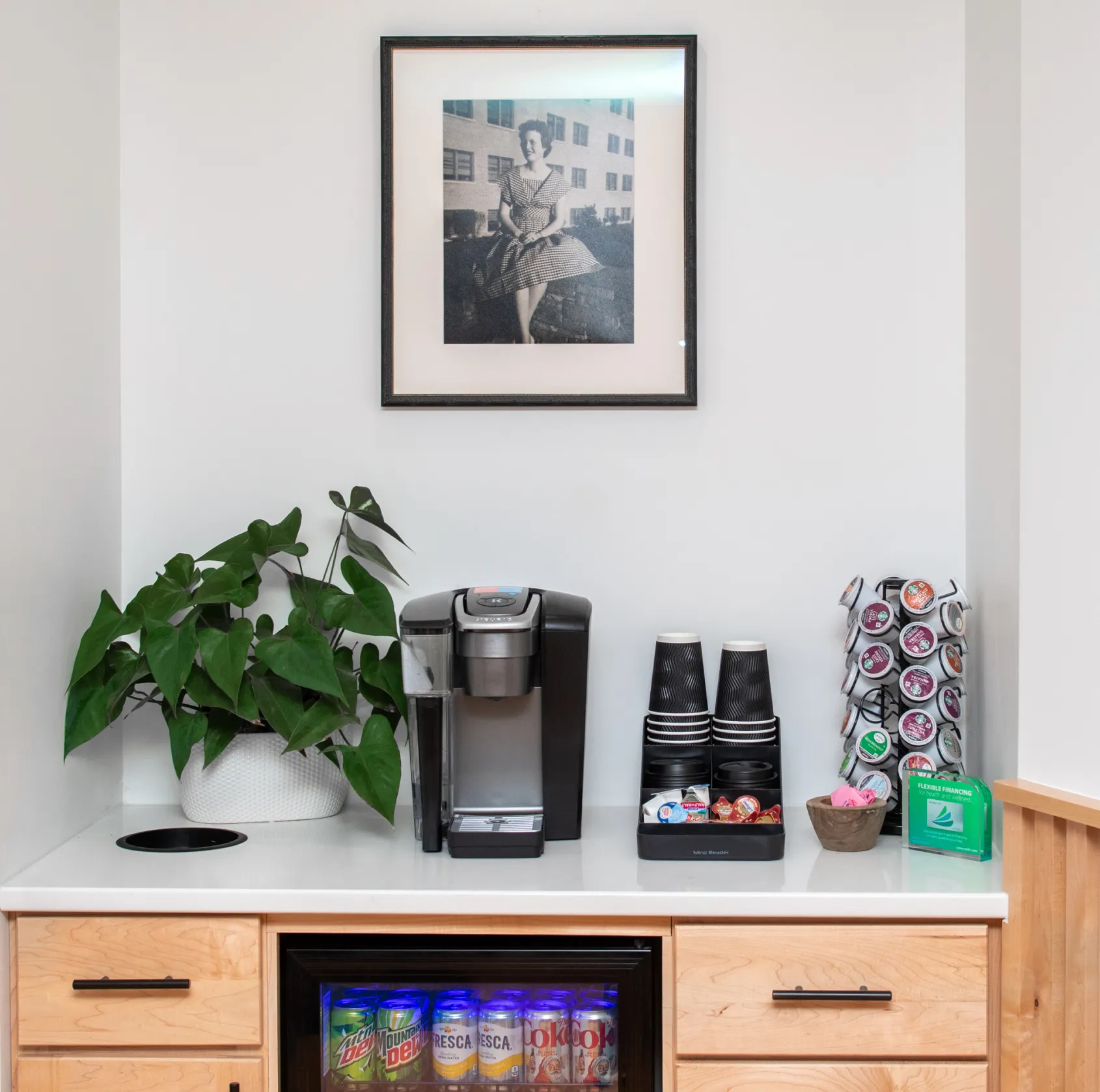 Coffee station with a coffee maker, disposable cups, coffee pods, a potted plant, and a mini fridge stocked with soda cans under a framed black-and-white photo of a woman sitting outdoors.