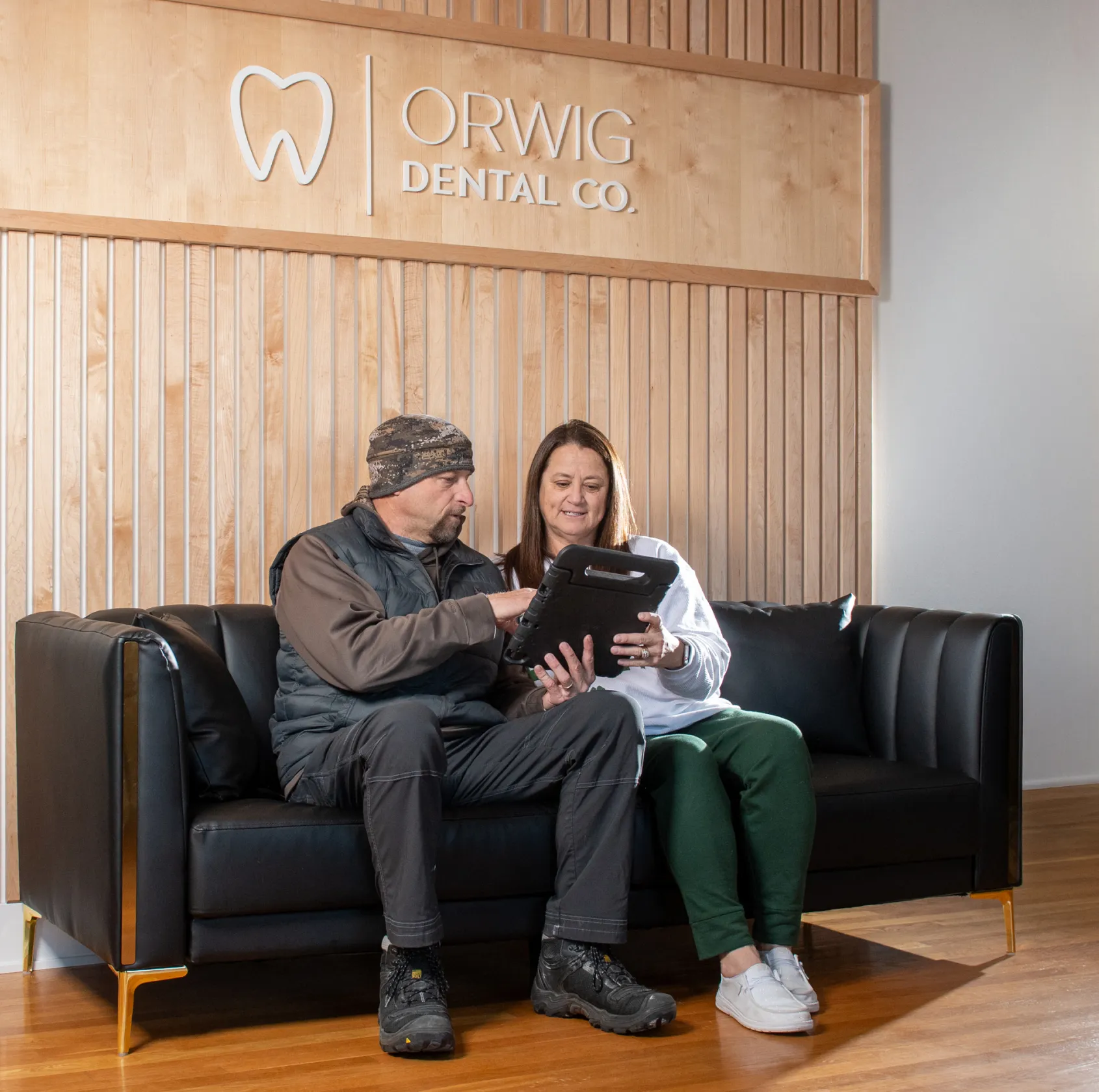 Two people sitting on a black leather couch in a dental office waiting area, looking at a tablet under the Orwig Dental Co. sign.