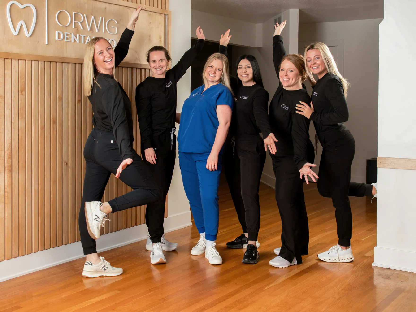 Six female dental staff members smiling and posing cheerfully in front of Orwig Dental Co. sign.
