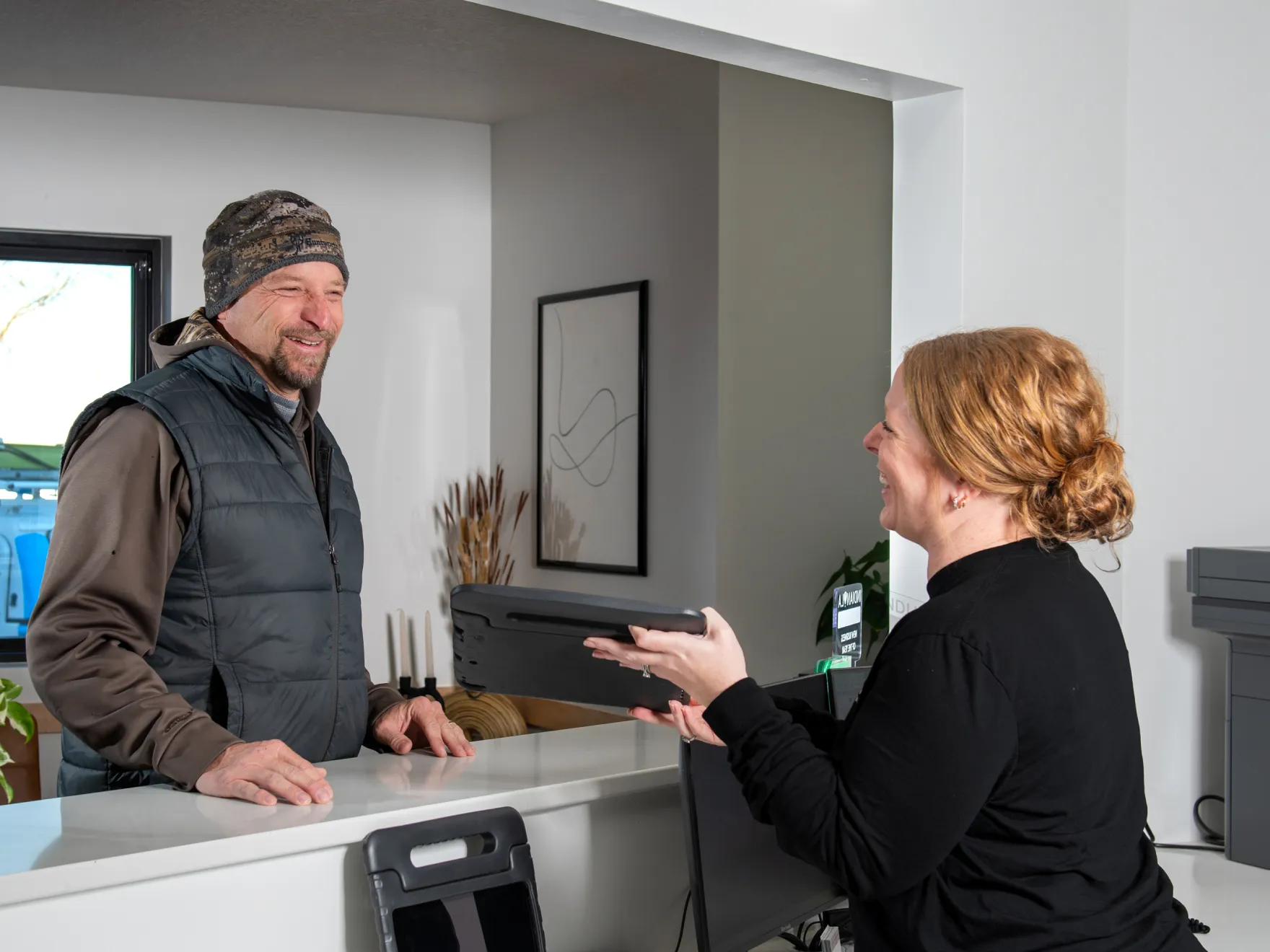 Smiling woman handing a tablet to a man wearing a hat and vest across a white counter in an office reception area.