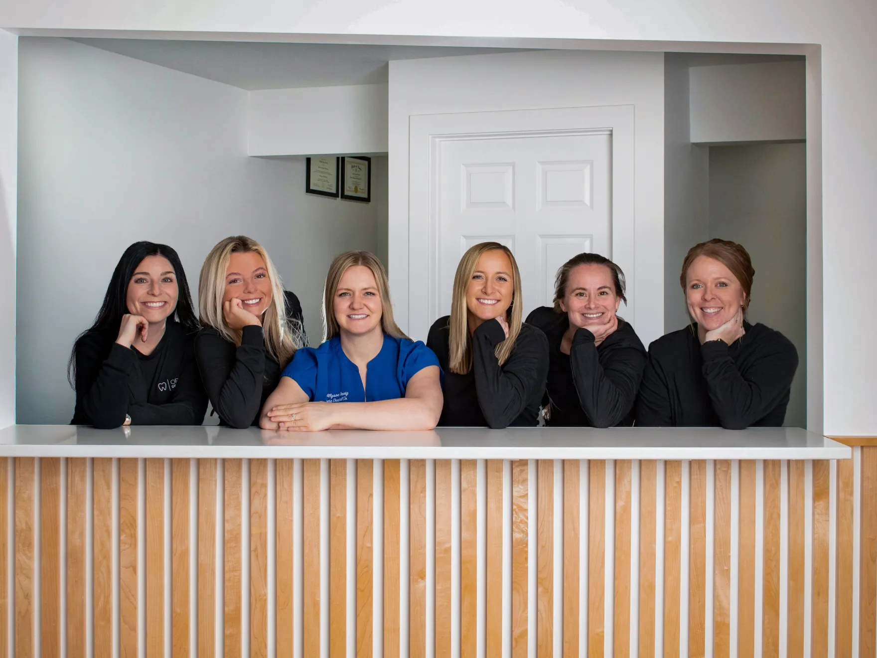 Six smiling women leaning on a white counter in a modern office setting.