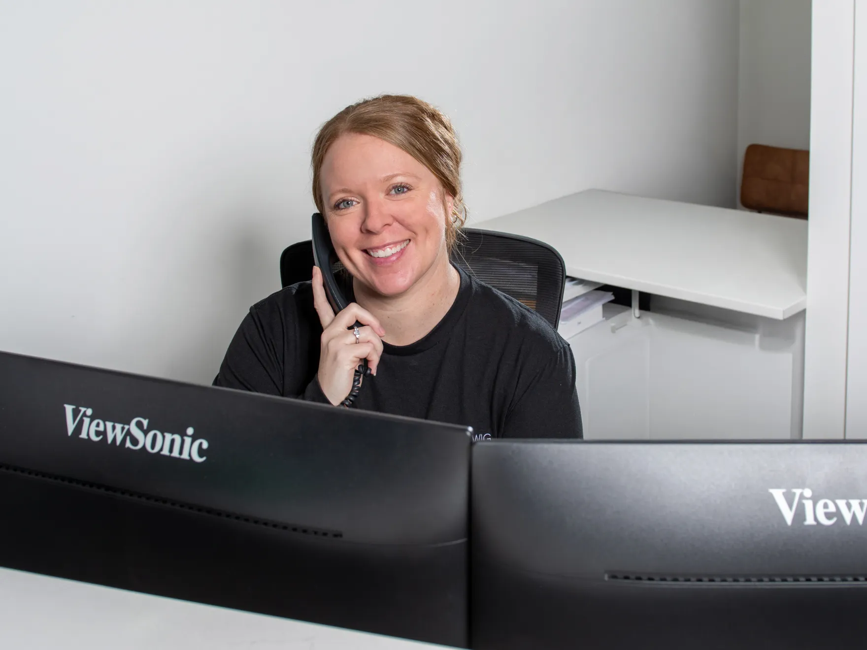 Smiling woman with light brown hair sitting at a desk and talking on a corded phone behind two ViewSonic monitors.