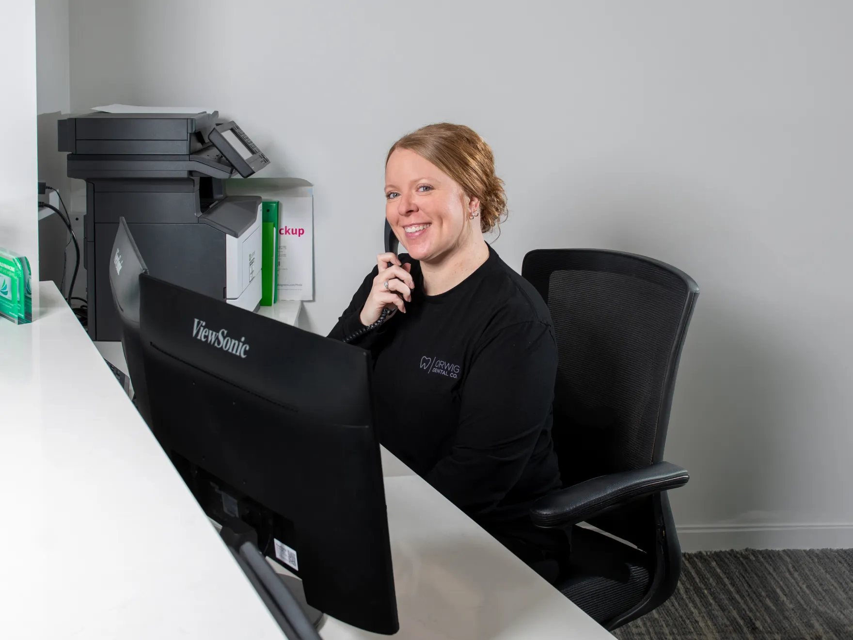 Smiling woman in black shirt sitting at a desk with dual monitors, talking on the phone in an office setting.