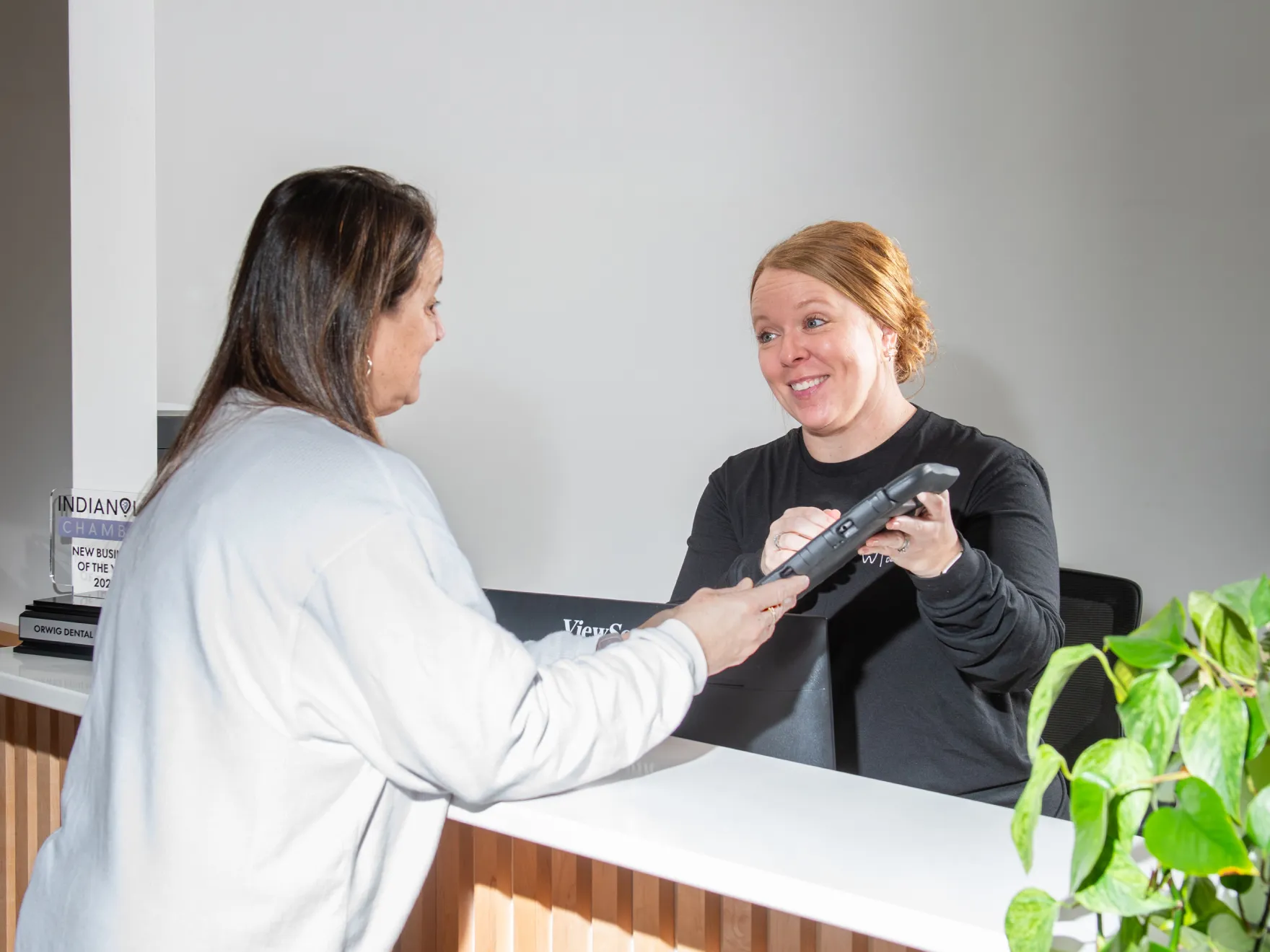 Receptionist smiling and handing a tablet to a woman standing at a desk in a modern office.