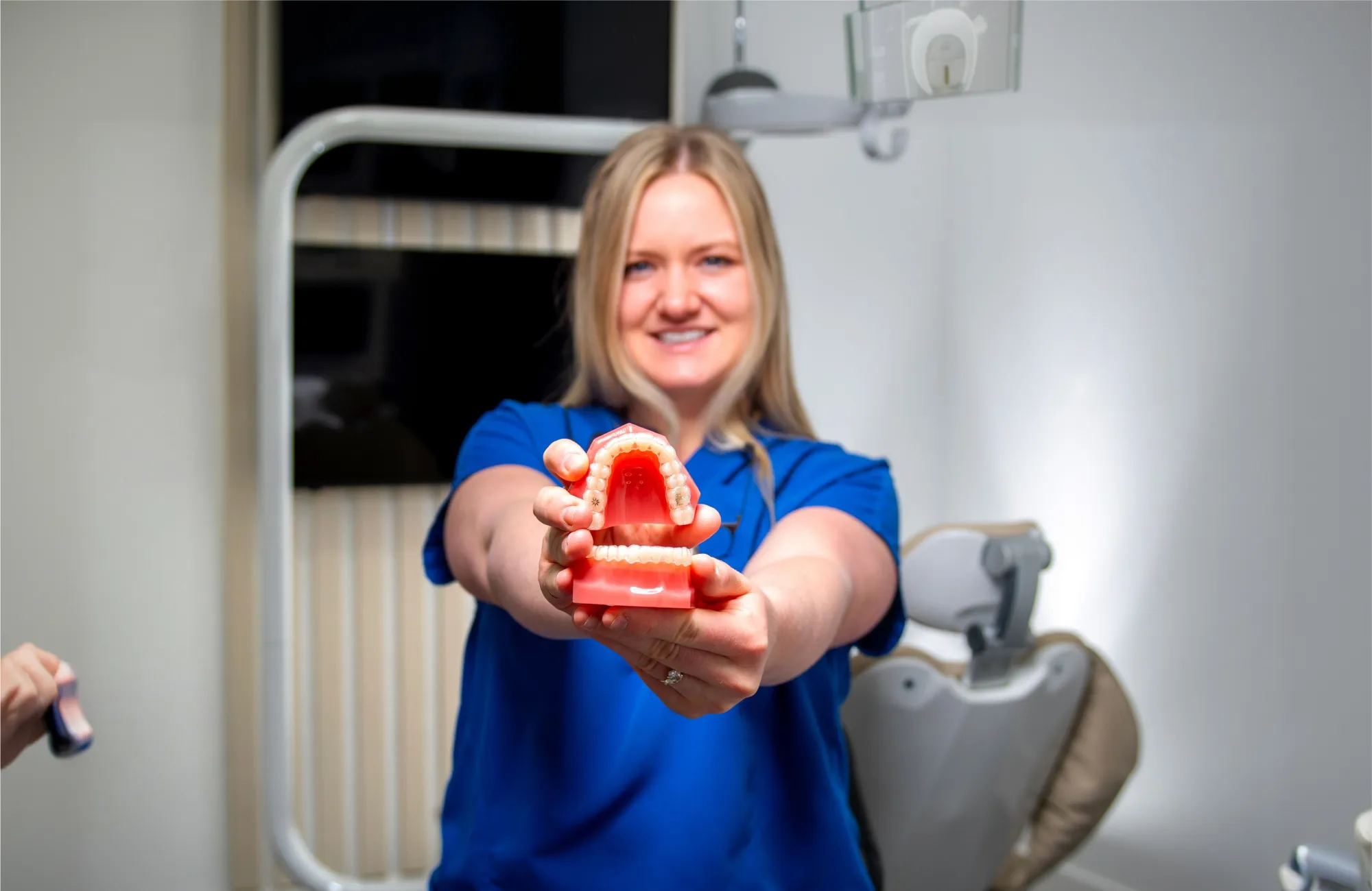 Smiling dental professional in blue scrubs holding a dental model showing upper and lower teeth inside a clinic.