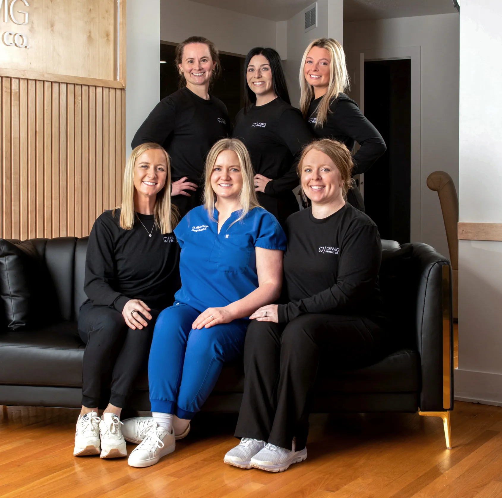 Group of six smiling women in medical attire sitting and standing around a black couch in a modern office setting.