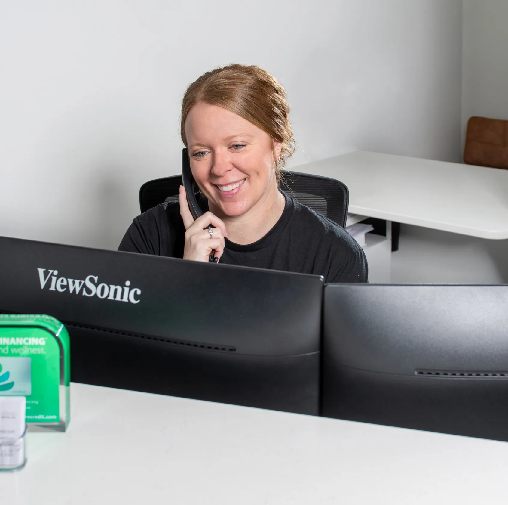 Smiling woman talking on a phone while sitting at a desk with dual ViewSonic monitors.