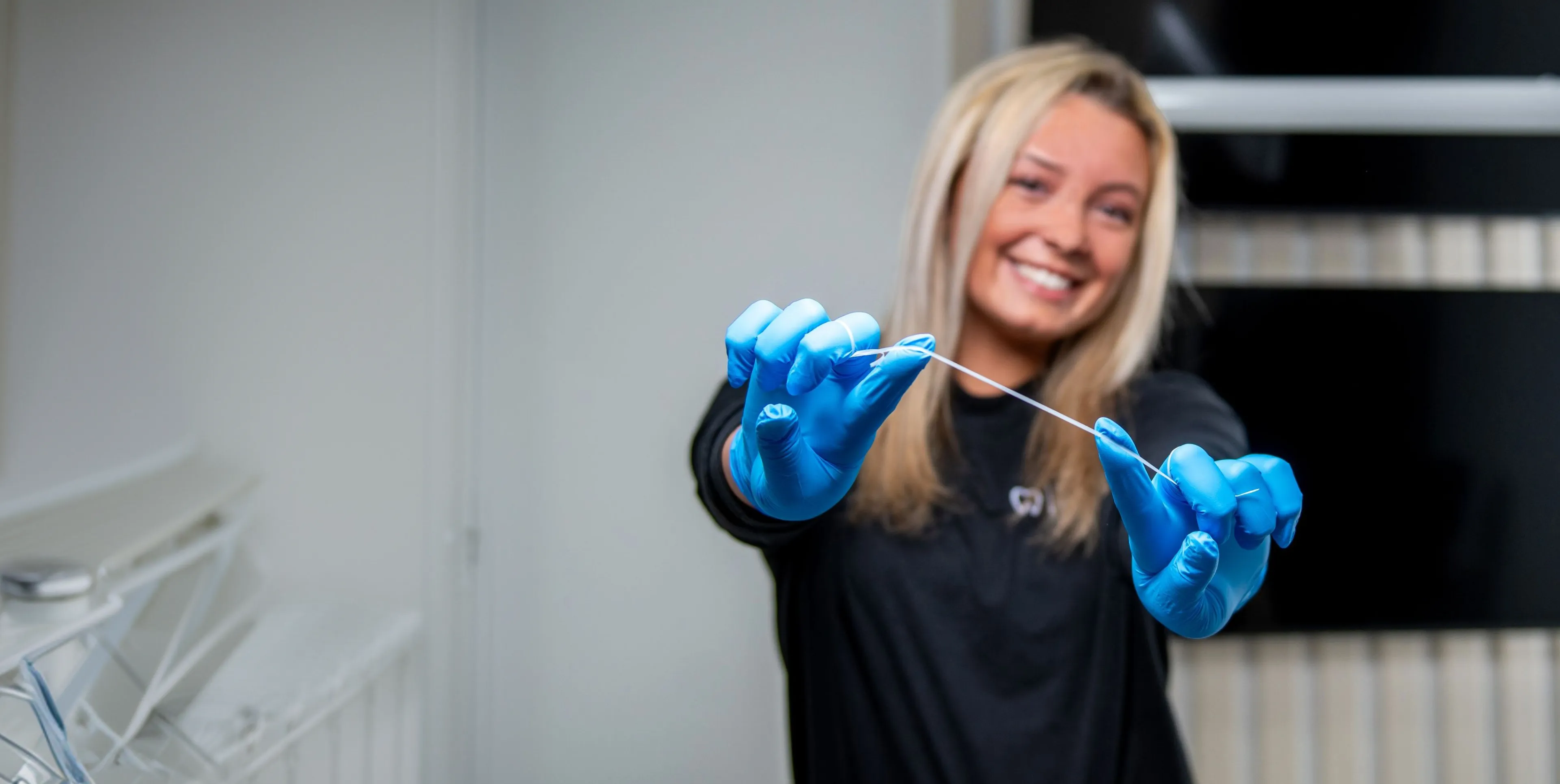 Smiling woman wearing blue gloves holding a stretched dental floss in a dental office.