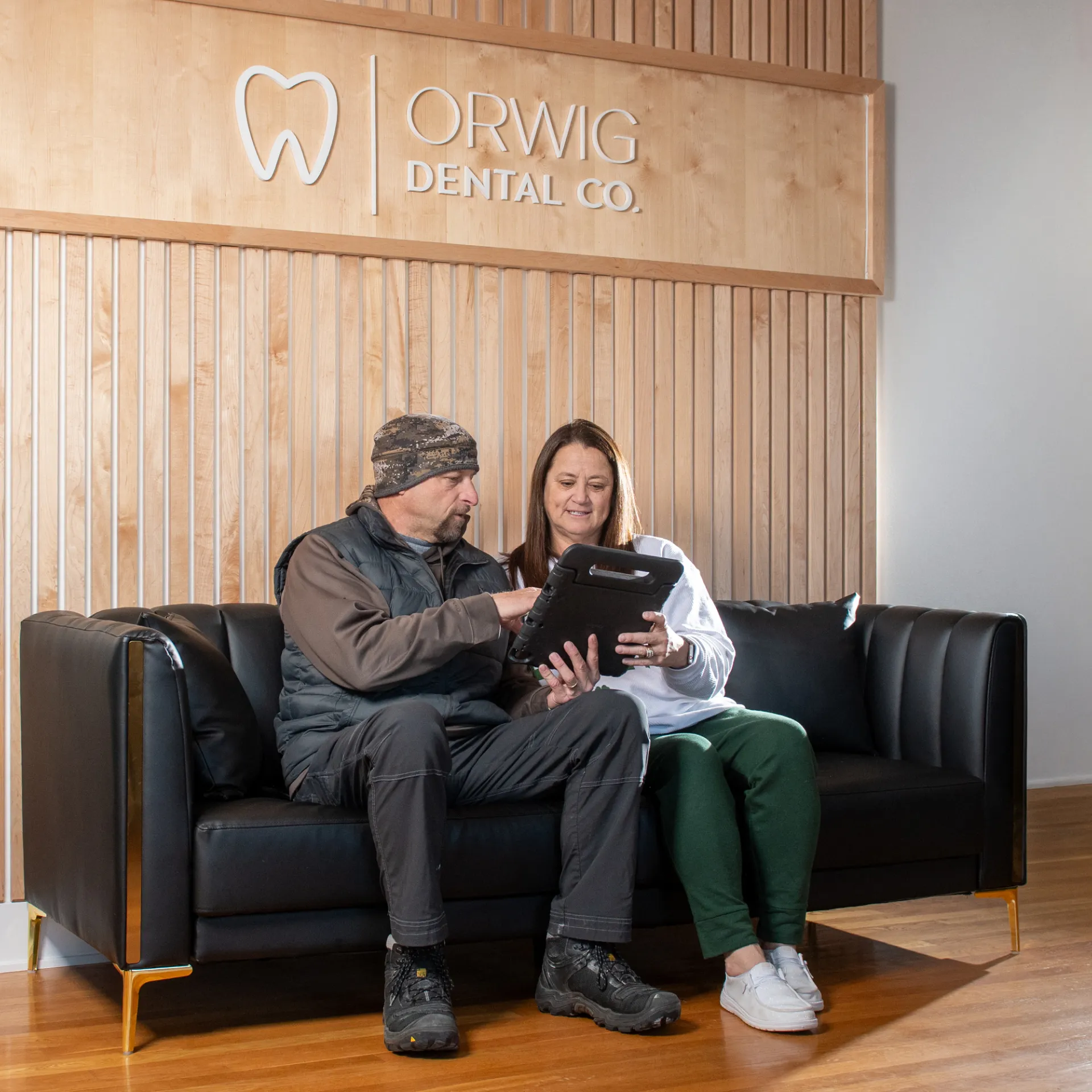 A man and a woman sitting on a black leather couch looking at a tablet in a dental office lobby with Orwig Dental Co. sign on a wooden panel wall.