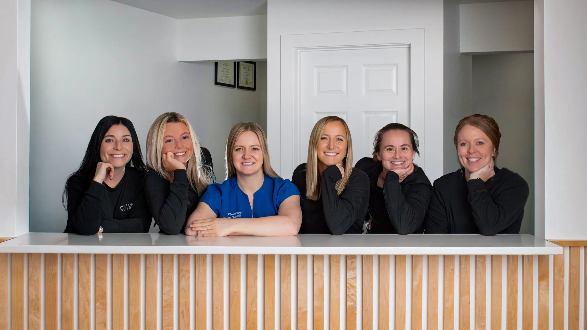 Six smiling women leaning on a white counter in a modern office setting, five wearing black tops and one in a blue top.