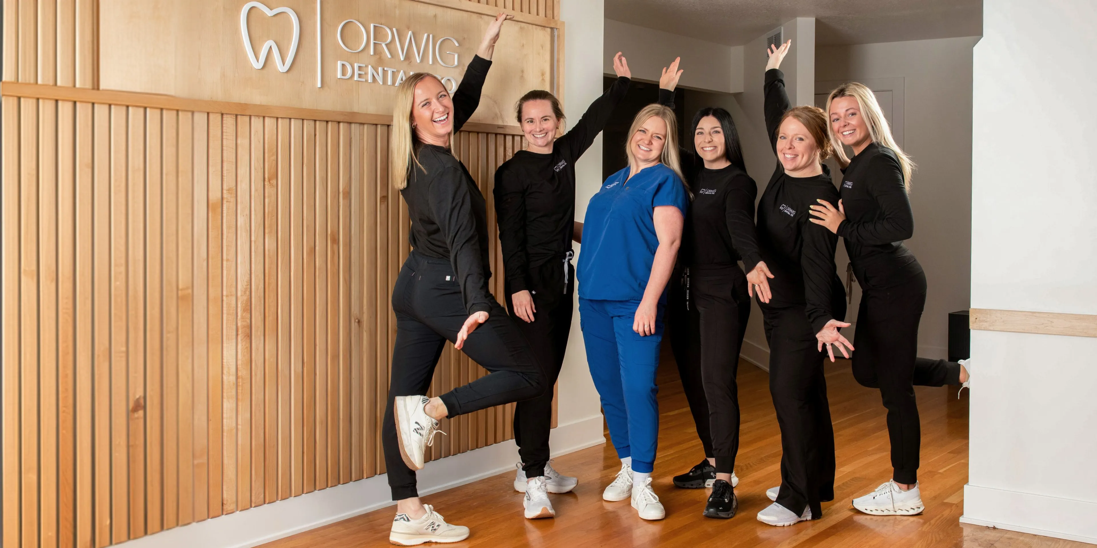 Six smiling dental staff posing playfully inside Orwig Dental office with wooden wall and logo background.
