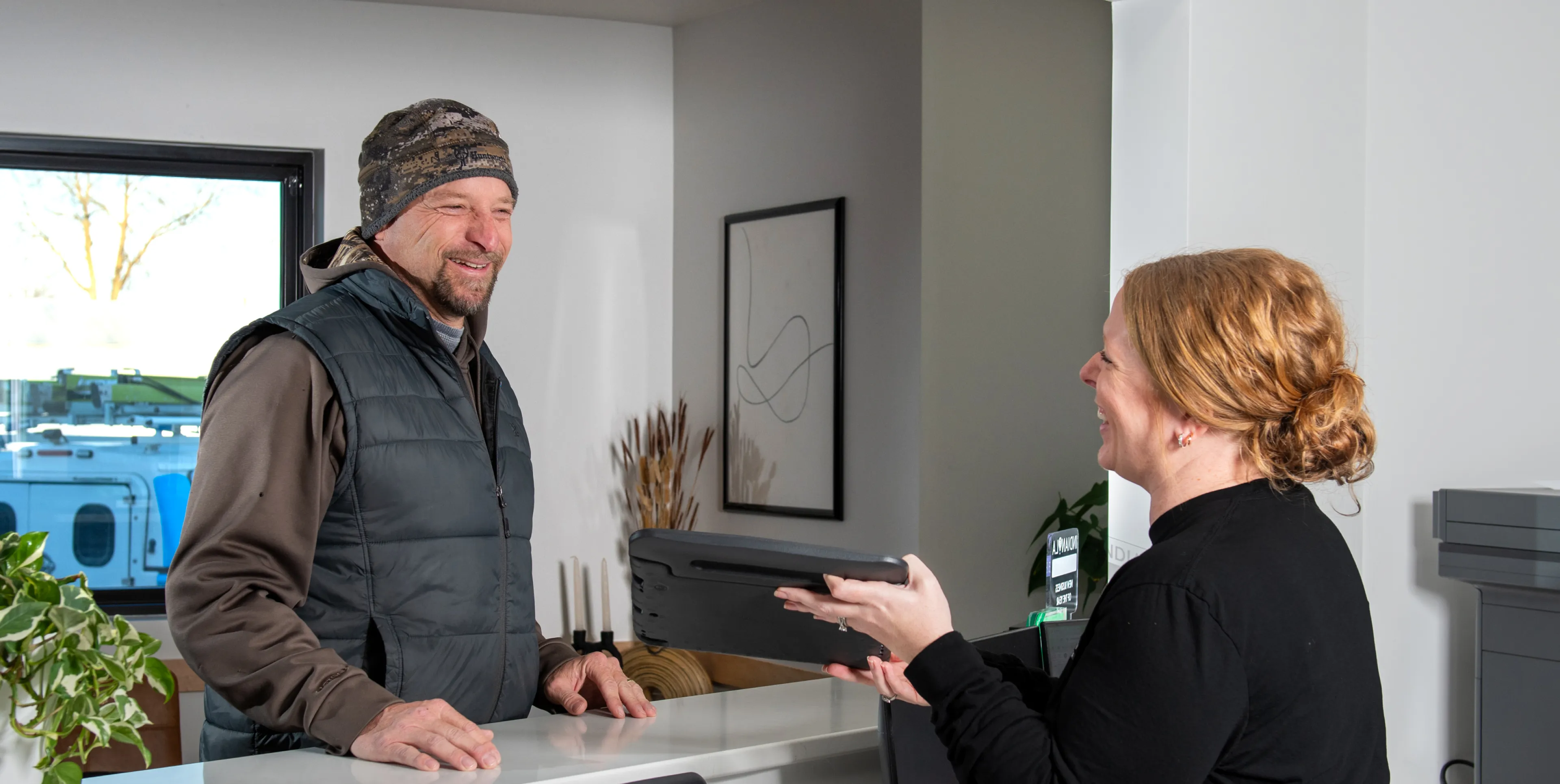 Man in a camouflage beanie and vest interacting with a smiling receptionist holding a tablet at a service counter.