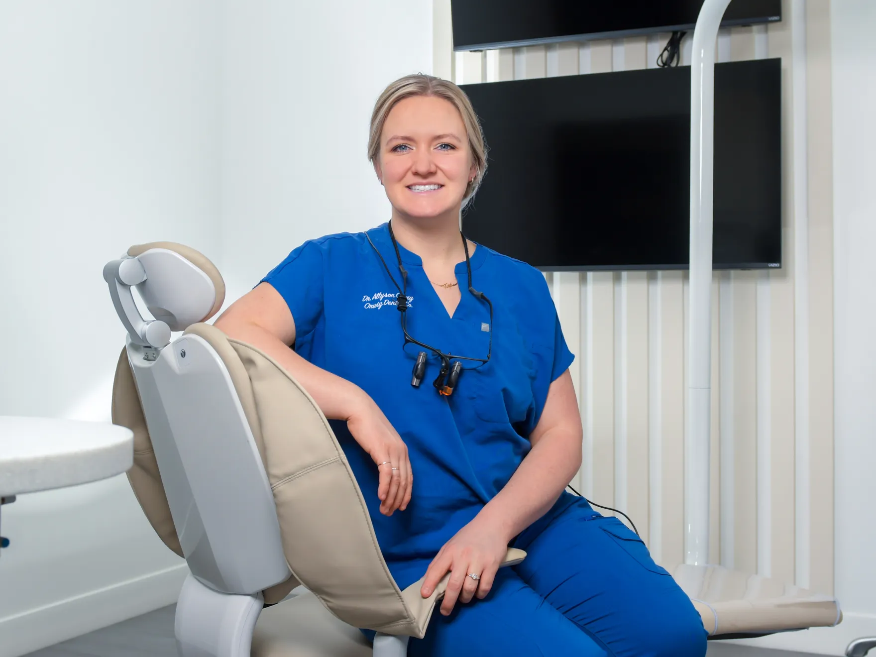 Smiling female dentist in blue scrubs sitting on a beige dental chair with dental loupes around her neck in a dental office.