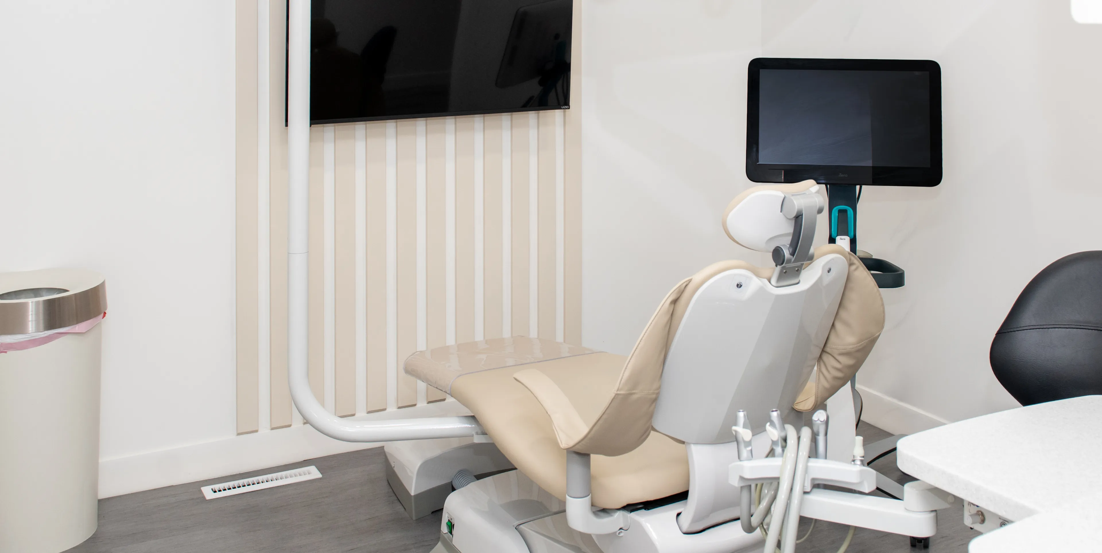 Dental office setup with beige dental chair, wall-mounted monitor, and dental instruments attached to the chair.