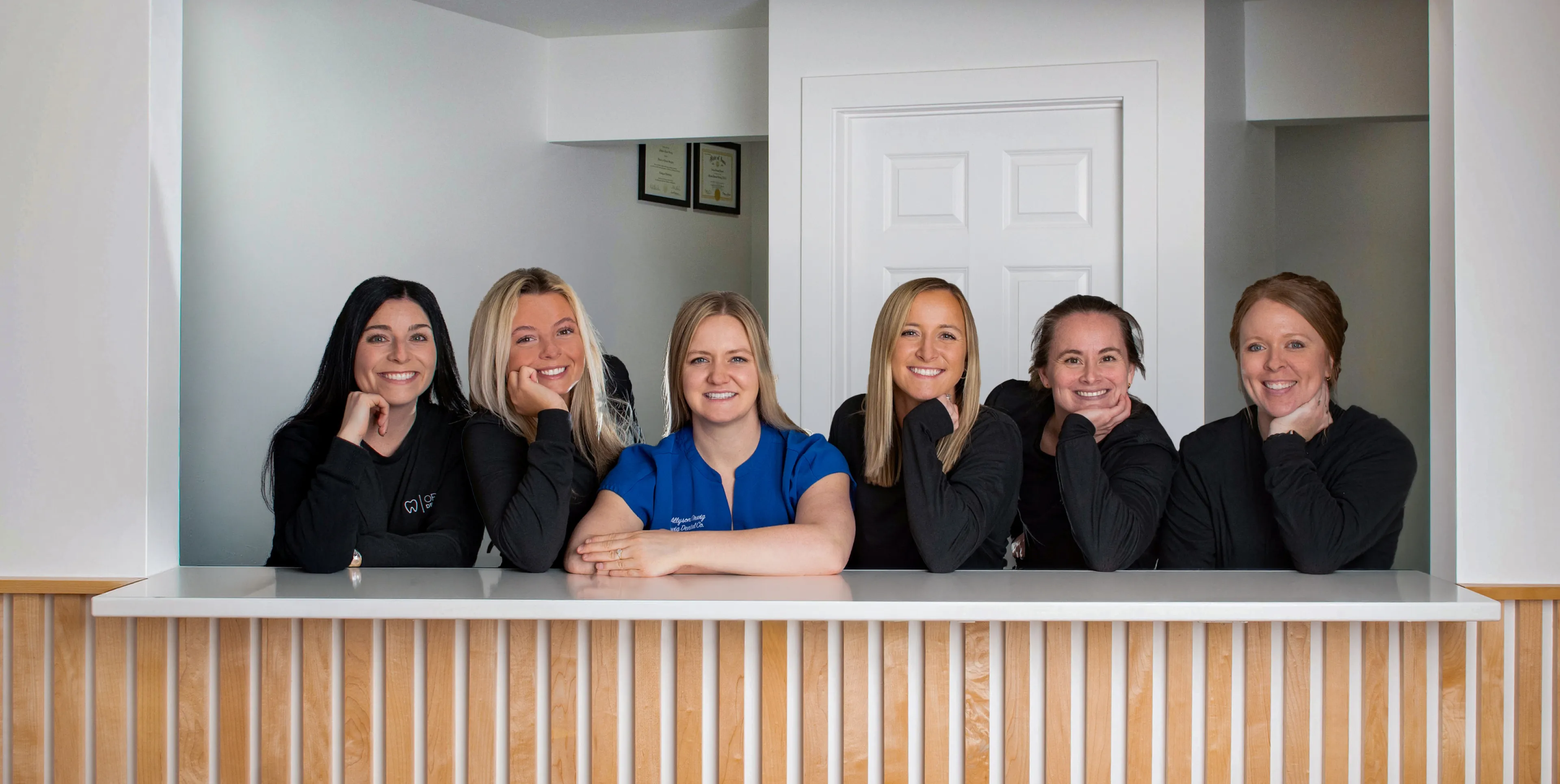 Six smiling women leaning on a white counter with a wooden slatted front, posing in a bright office space.