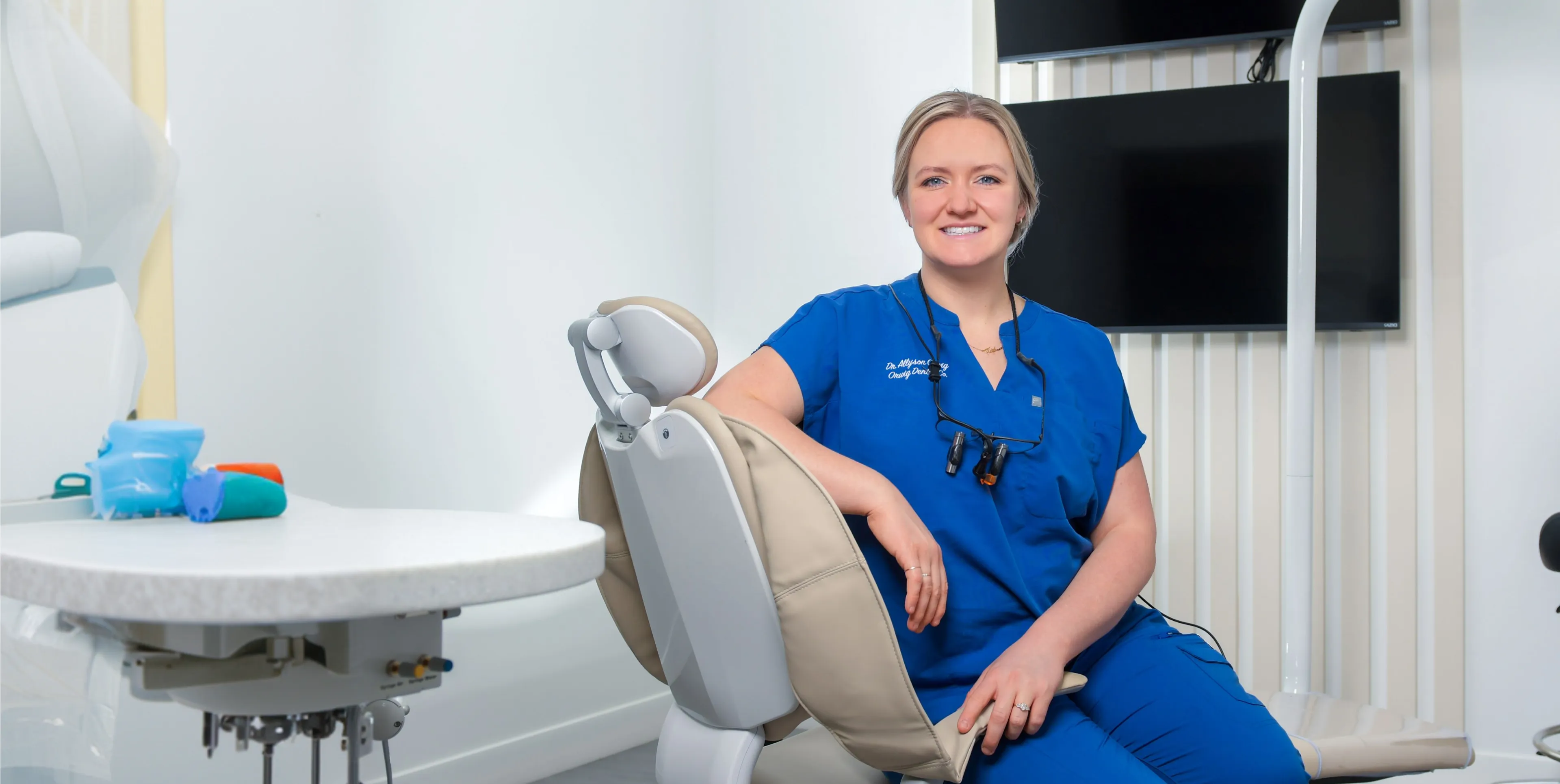 Smiling female dentist in blue scrubs sitting on a beige dental chair in a bright dental office.
