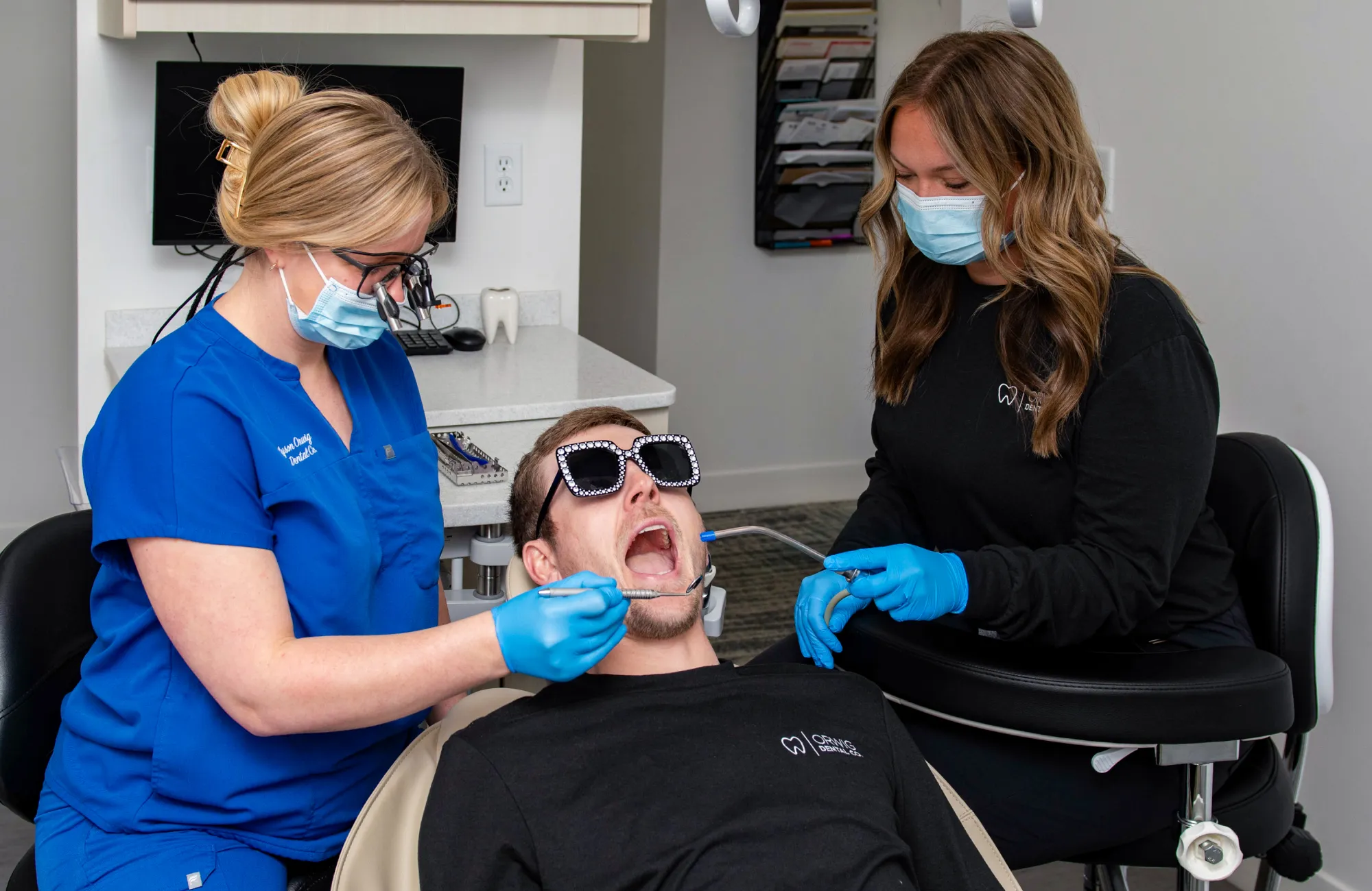 Dentist and dental assistant wearing masks and gloves treating a male patient reclining in a dental chair with sunglasses on.