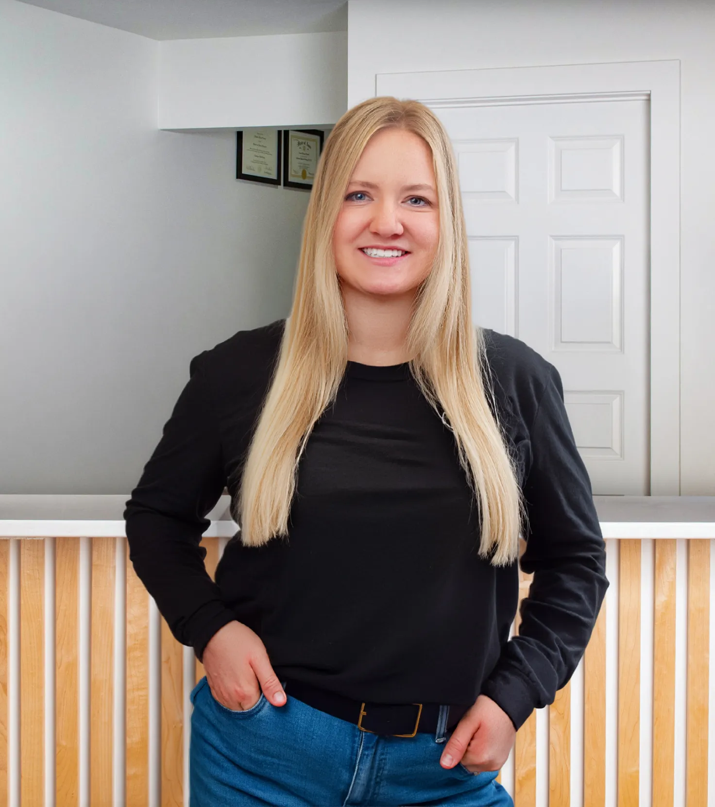 Smiling woman with long blonde hair wearing a black long-sleeve shirt and blue jeans standing indoors with hands in pockets.