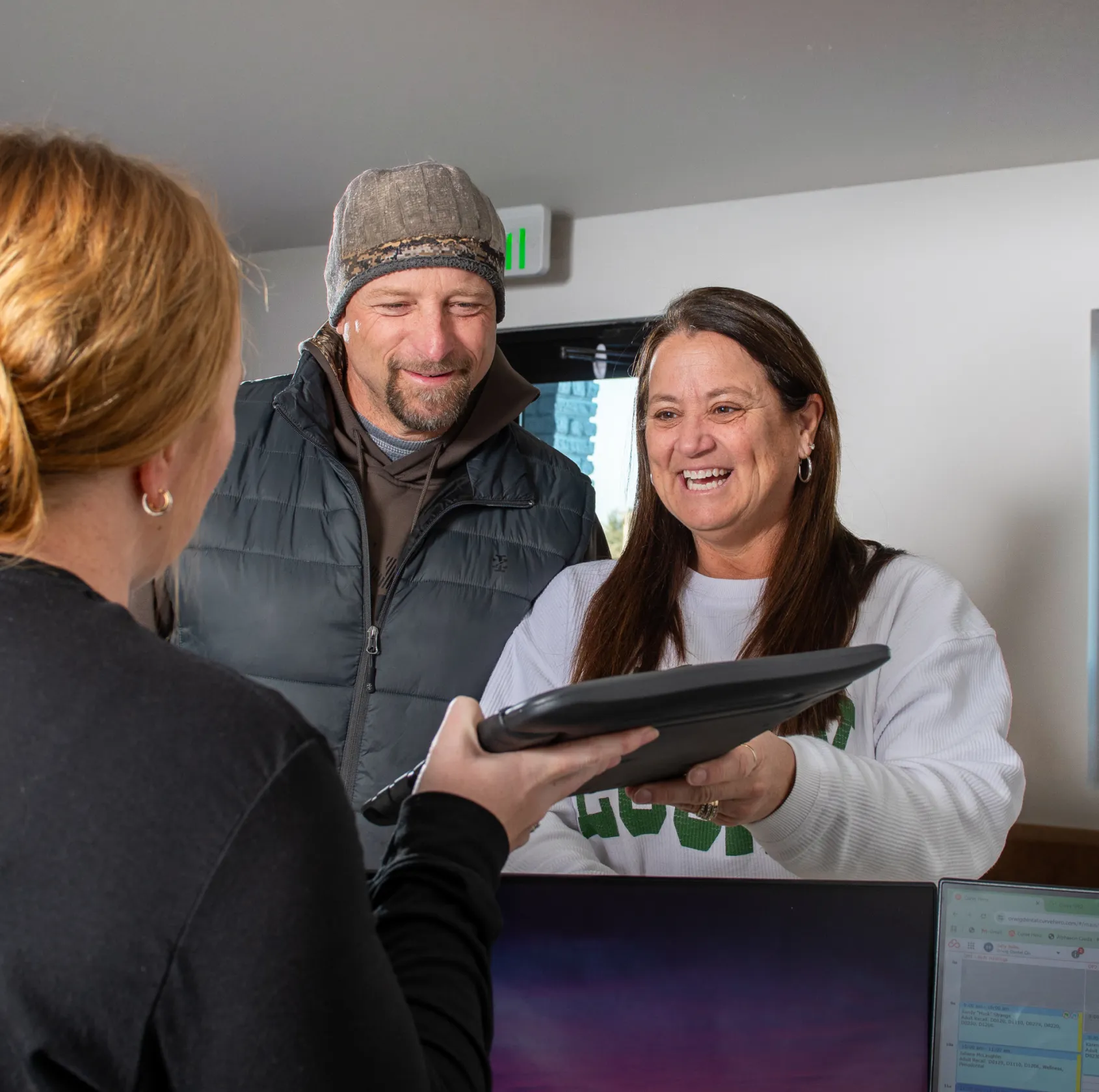 Smiling woman receiving a black folder from another person across a reception desk with a man standing beside her.