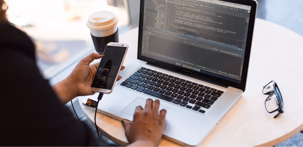 Image of laptop with code on it and a person looking at their cell phone.