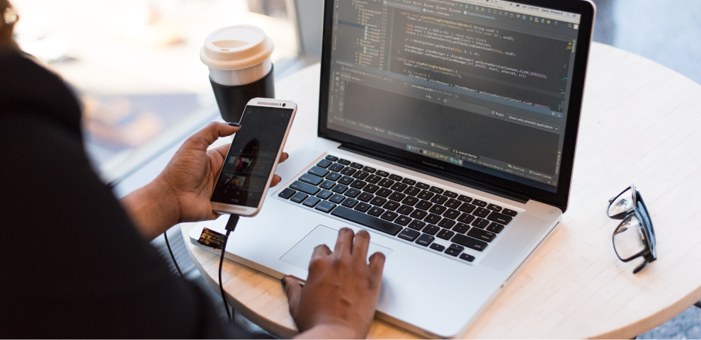 Image of laptop with code on it and a person looking at their cell phone.