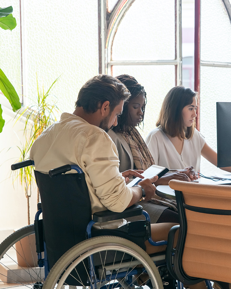 Boss in wheelchair reviews ipad while in meeting with two coworkers at a copmuter.