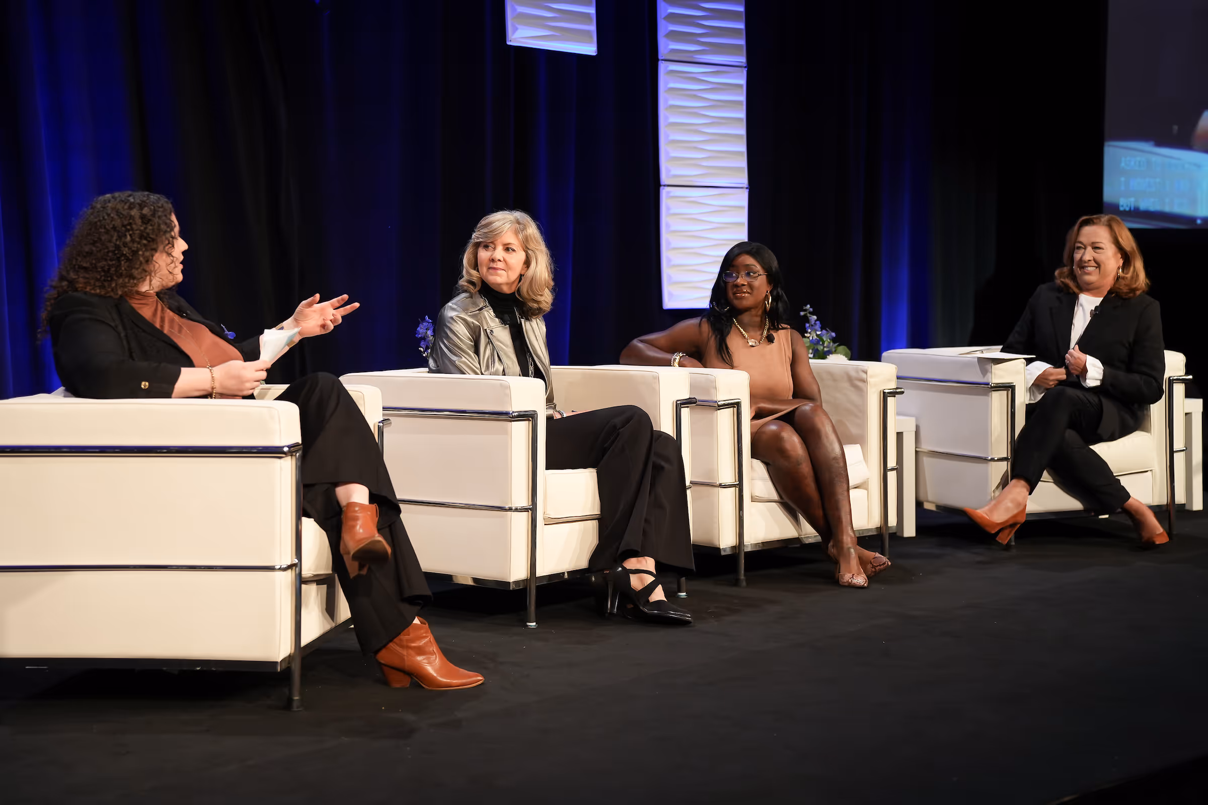 Four women seated on white armchairs on a stage engaged in a panel discussion.