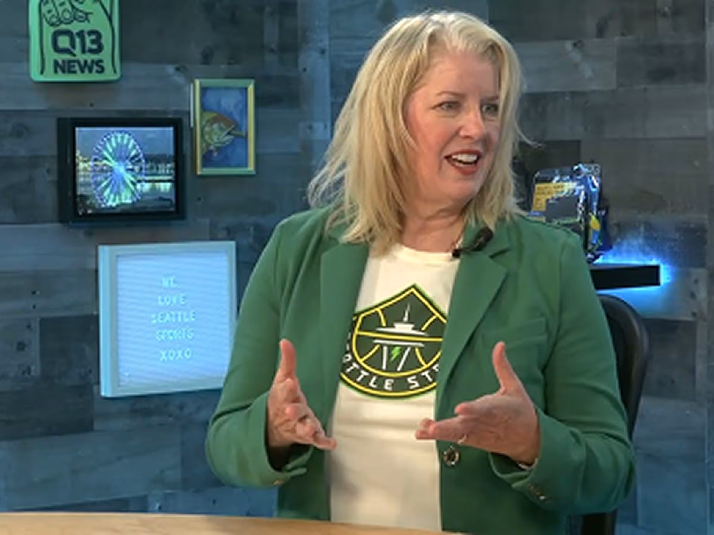 Woman in green blazer talking and gesturing, with Seattle-themed decor in background including Q13 News sign.