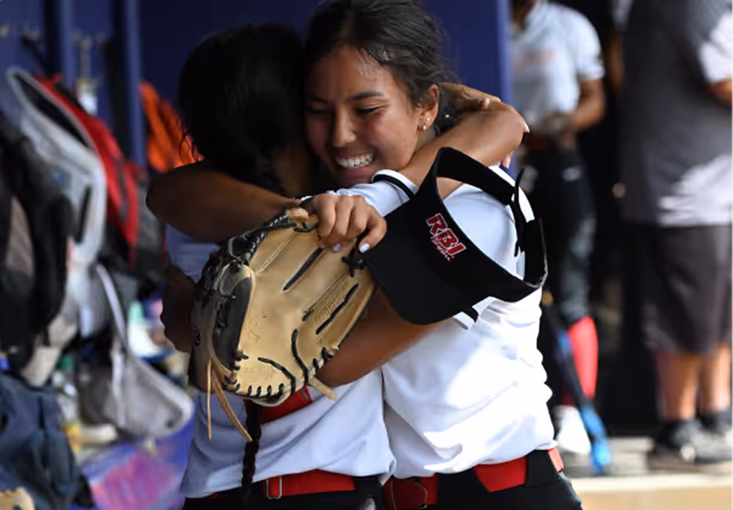 Two female softball players hugging, one holding a glove and a black RBI visor, smiling happily.