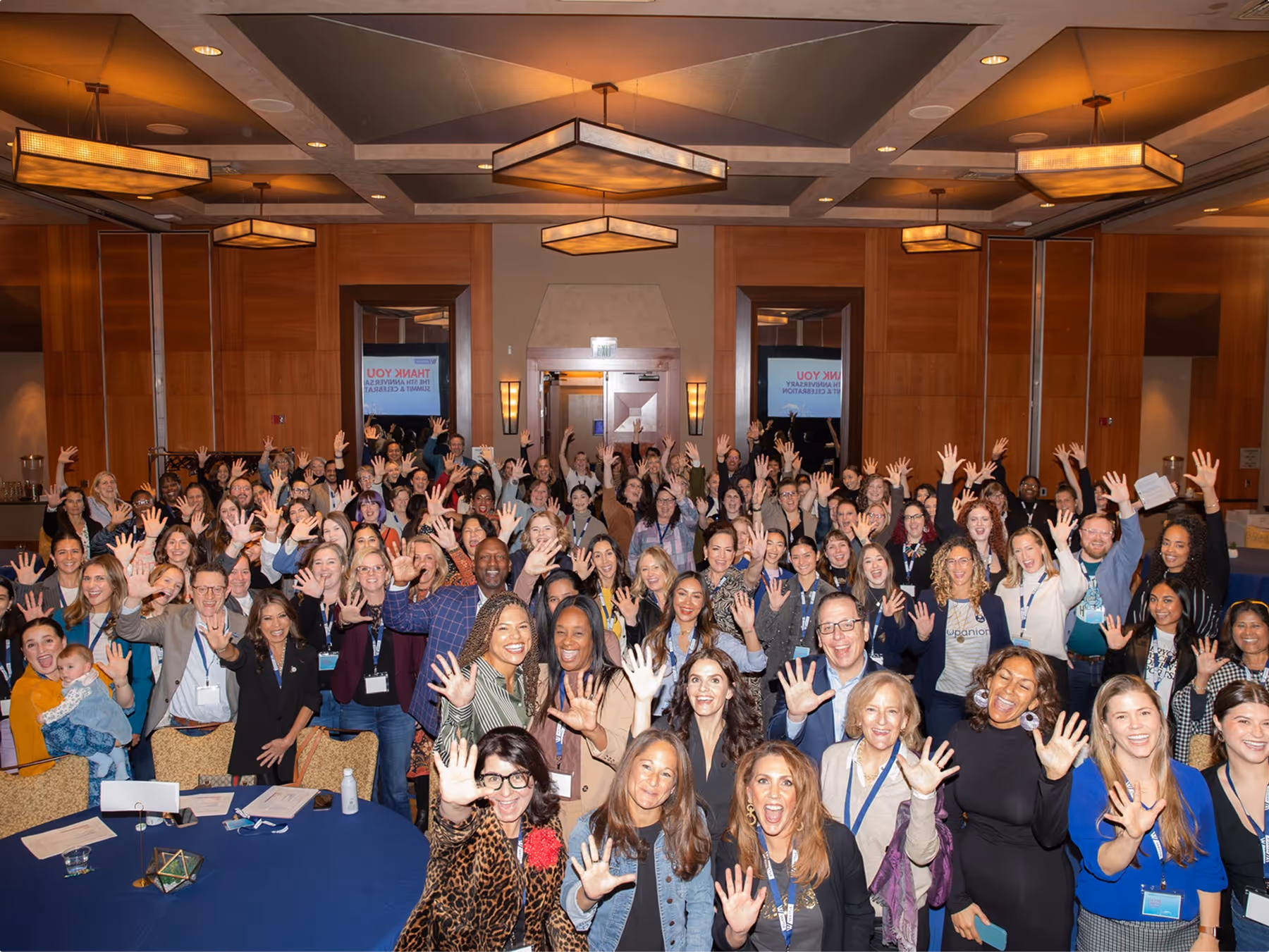 Large diverse group smiling and raising hands in a well-lit conference room.
