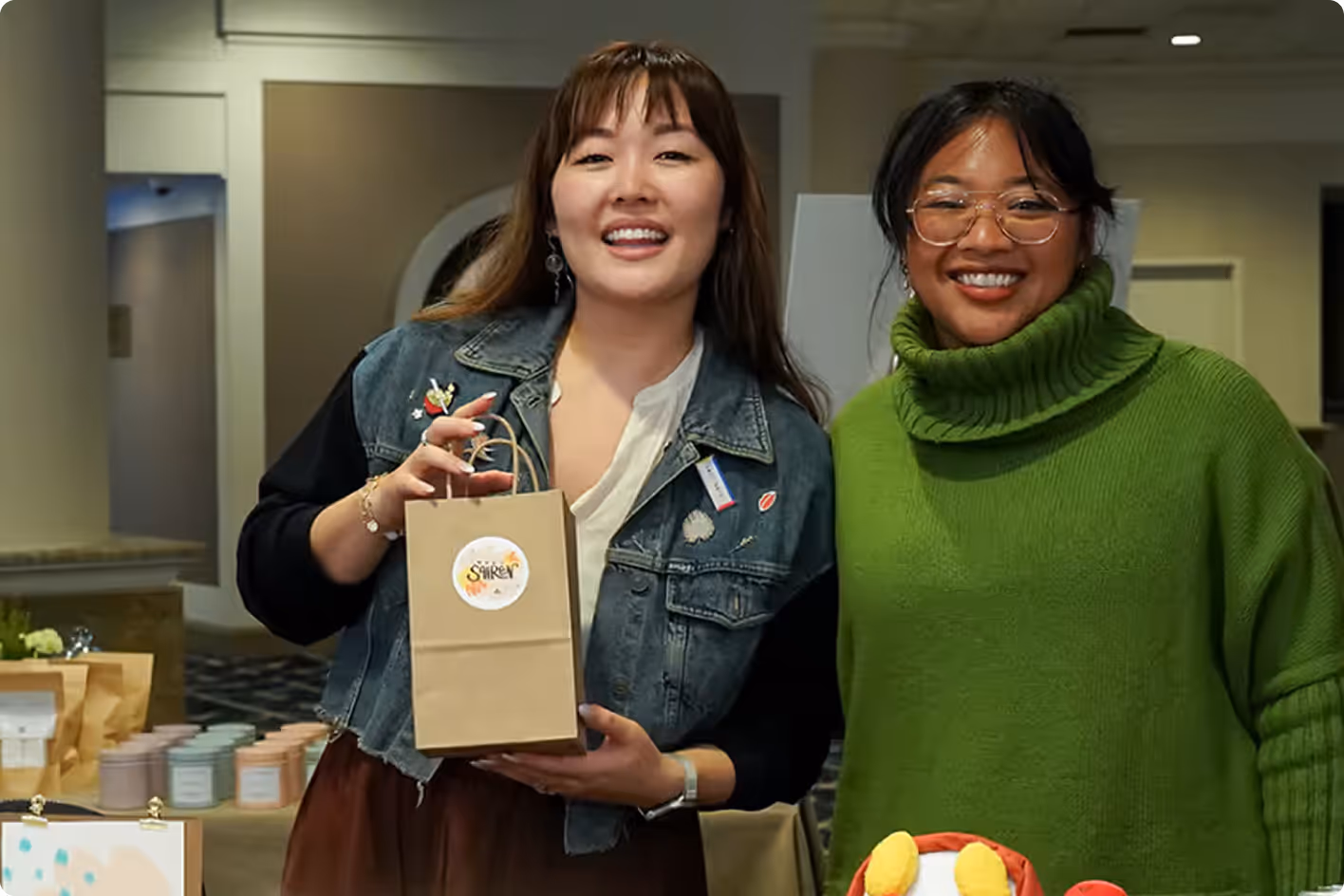 Two women smiling indoors; one holds a brown paper bag with a 'SIREN' logo sticker.