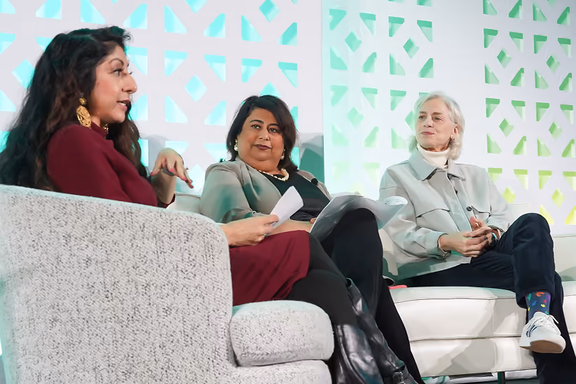 Three women seated on stage during a discussion or panel, with one woman speaking while holding papers and the other two listening attentively.