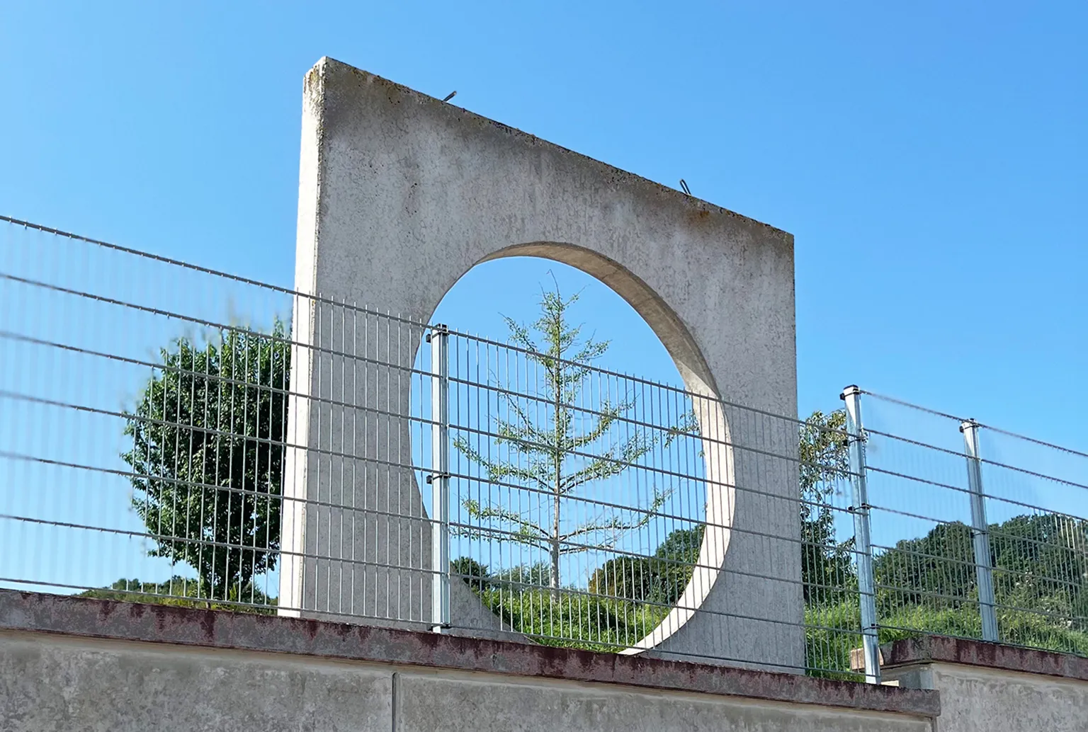 Betonmauer mit rundem Durchbruch, Baum sichtbar, Zaun und Landschaft im Hintergrund