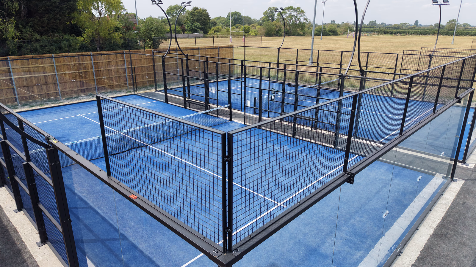 Two empty blue paddle tennis courts enclosed by black metal fences and transparent glass panels, with outdoor lights and a field visible in the background.