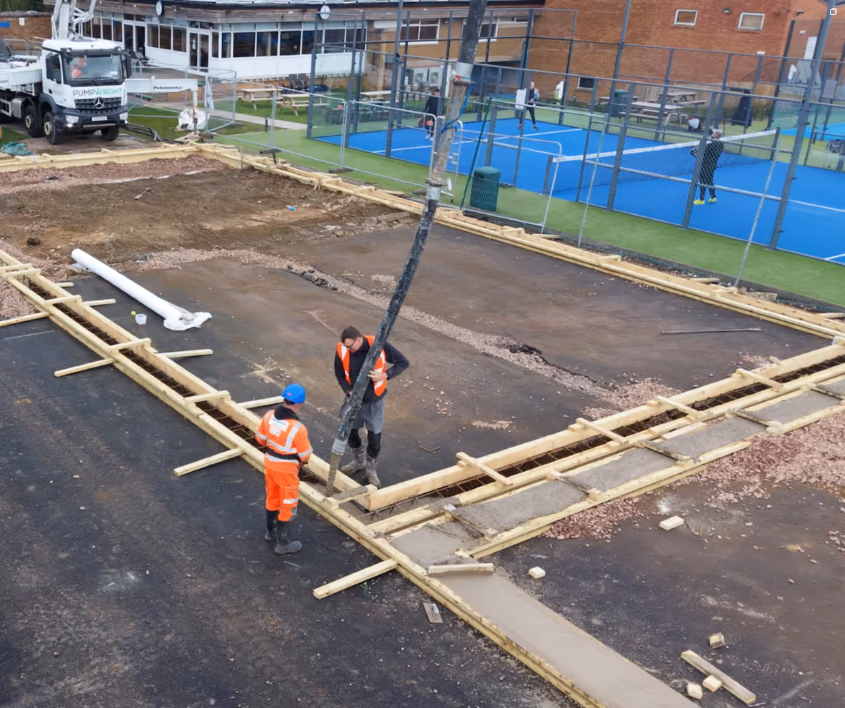 Two construction workers in safety vests and helmets working on pouring concrete within wooden frames at a construction site with padel tennis courts and a truck in the background.