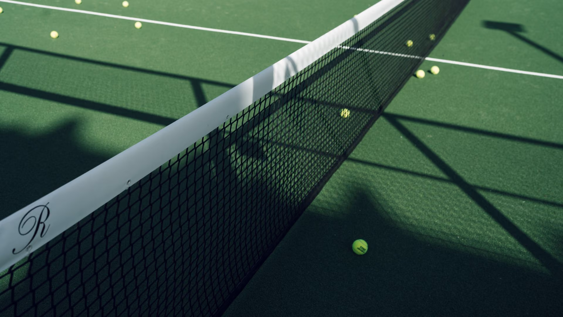Close-up view of a tennis net on a green court scattered with several yellow tennis balls under sunlight.