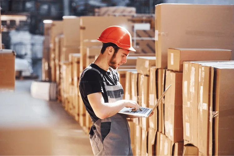 Storage worker in uniform and modern laptop in hands checks production