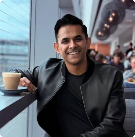 Smiling man in a black leather jacket sitting by a window with a cup of coffee in a cafe.