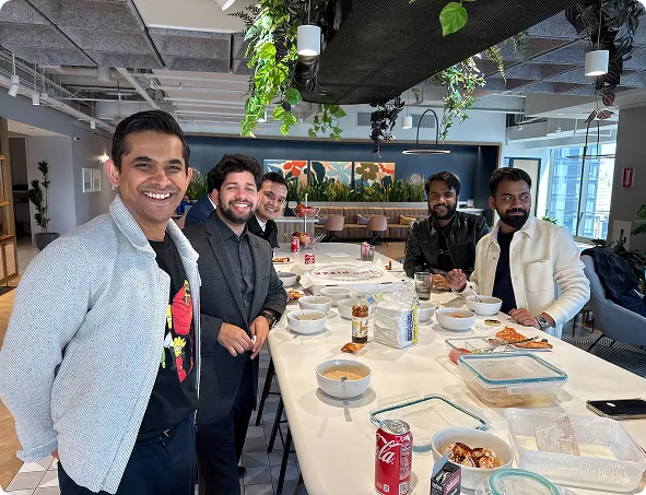 Five men sitting and standing around a table with bowls, food containers, and soda cans in a modern office.
