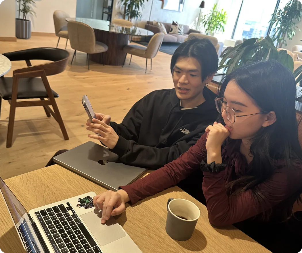 Two people collaborating on a laptop at a table, one using phone, with coffee cup nearby in a modern room.