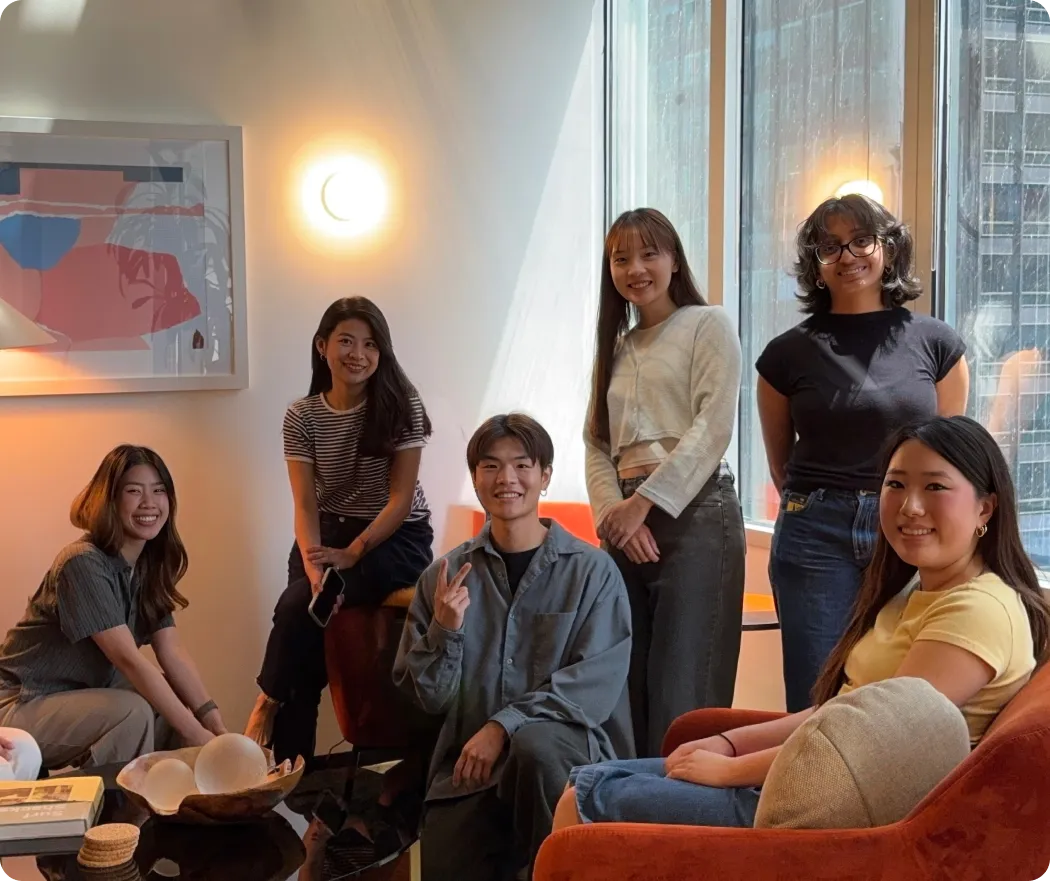 Group of six young adults smiling, sitting and standing in a cozy room with large windows and modern decor.