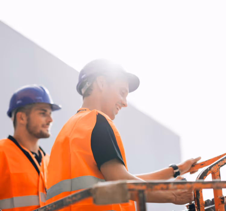 Two construction workers wearing orange safety vests and helmets operating machinery outdoors.