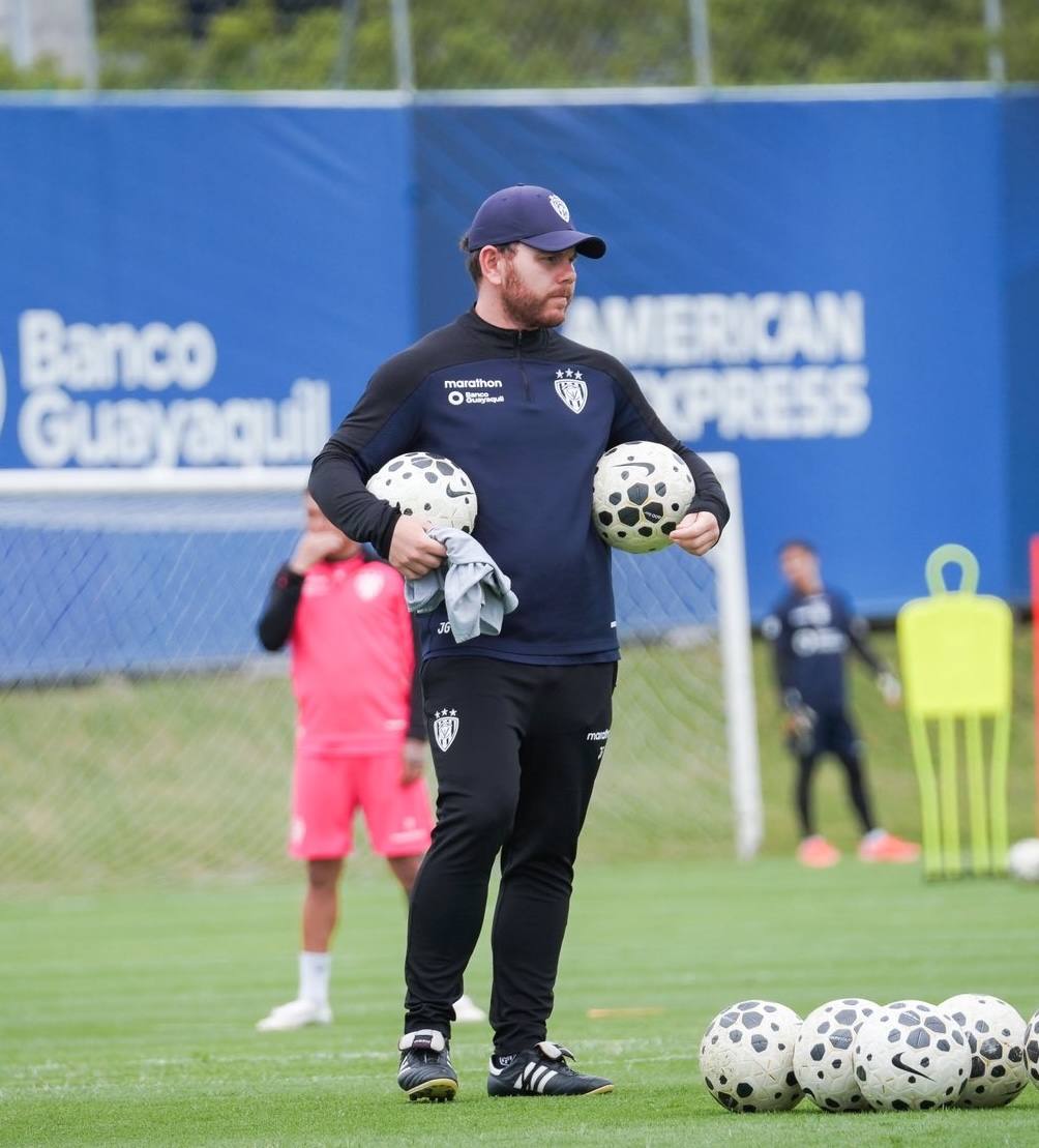 Man with a beard wearing white earbuds and a black shirt, sitting in front of a dark wooden bookshelf filled with books and decorative items.