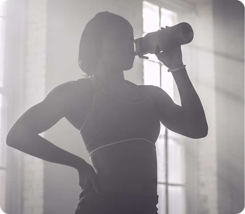 Silhouette of a woman in athletic wear drinking from a bottle with light streaming from windows.