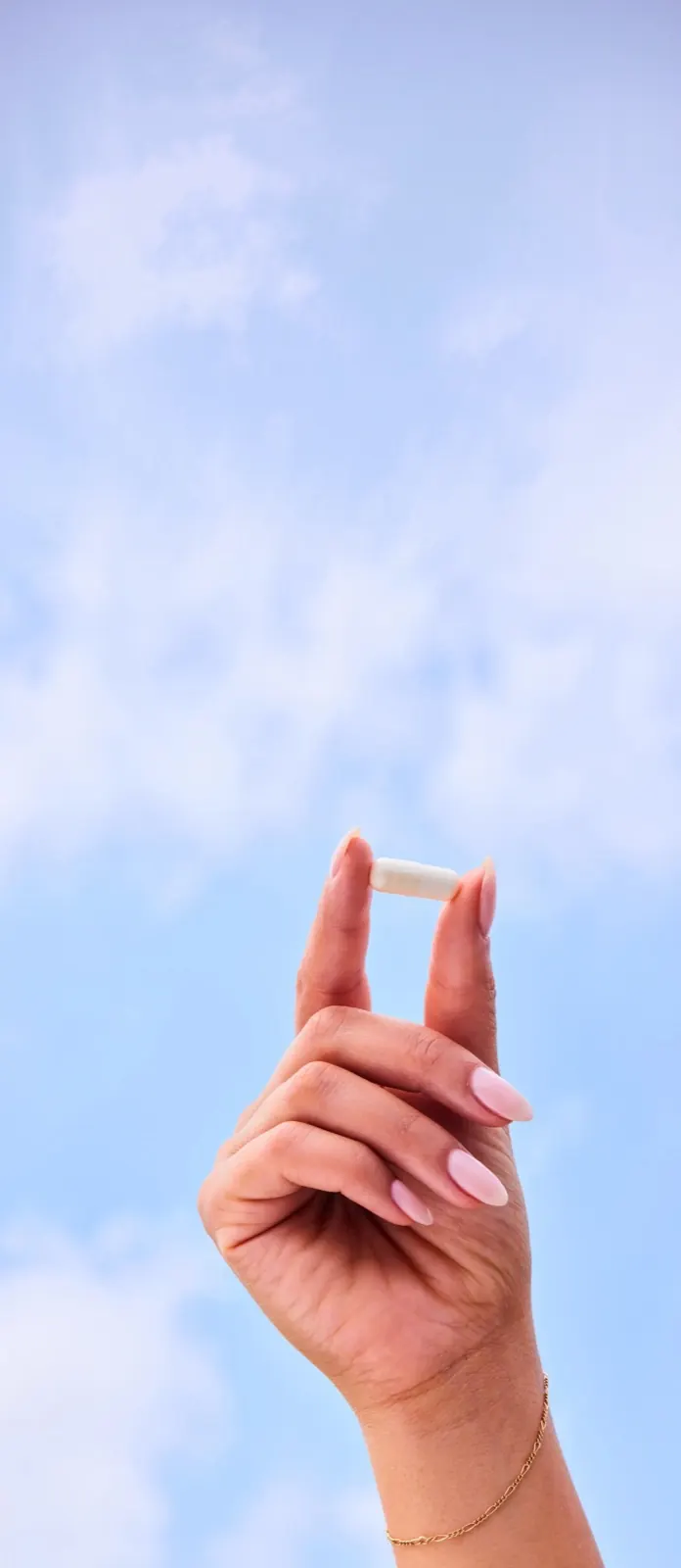Hand holding a single supplement capsule against a bright blue sky