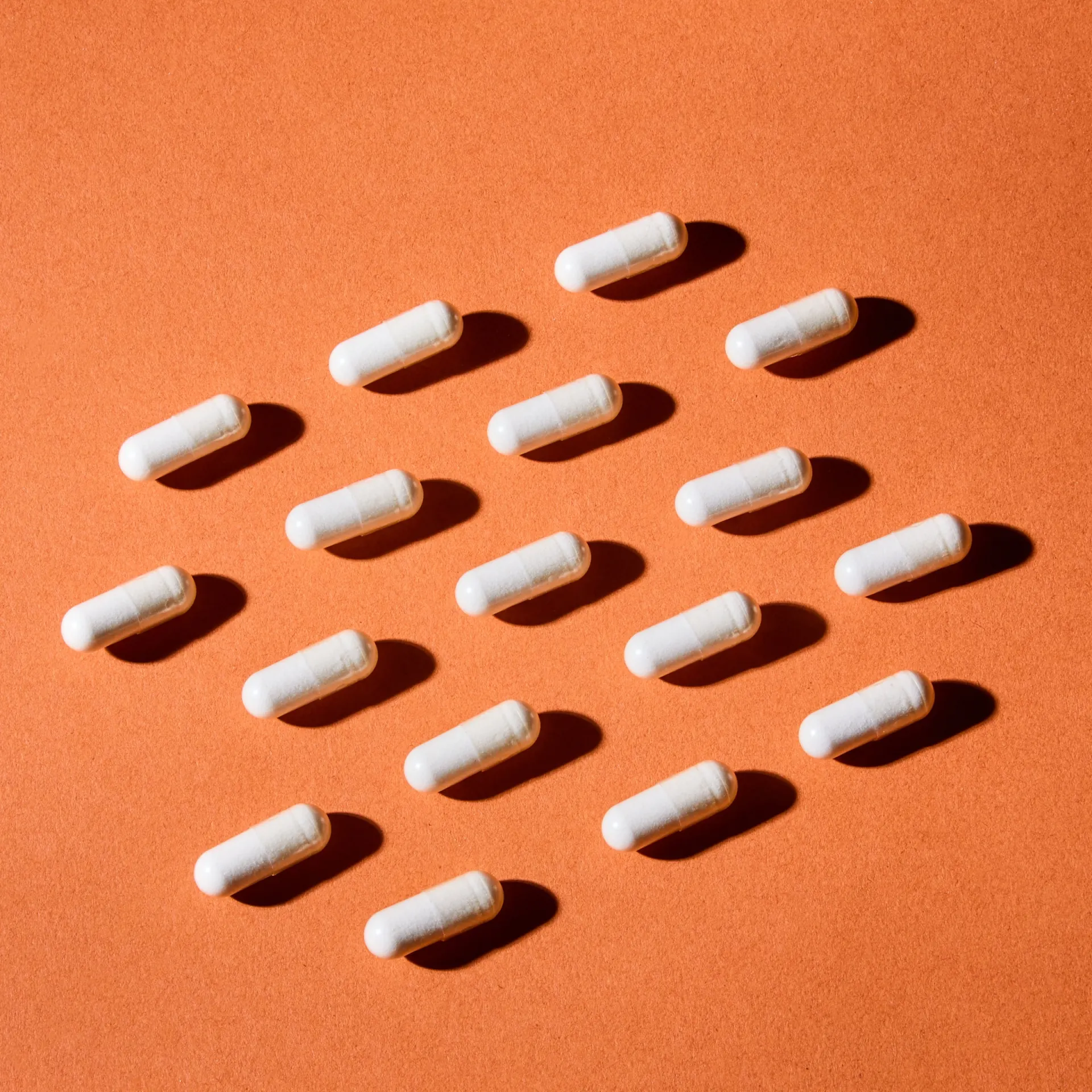Rows of white supplement capsules casting shadows on an orange surface