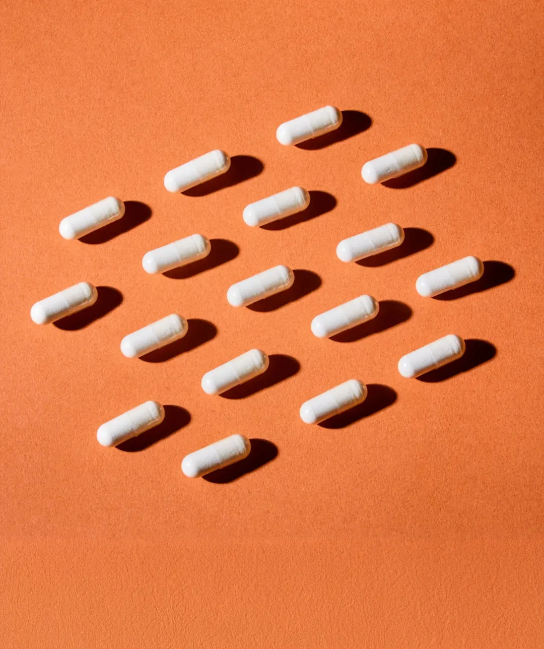 Rows of white supplement capsules casting shadows on an orange surface