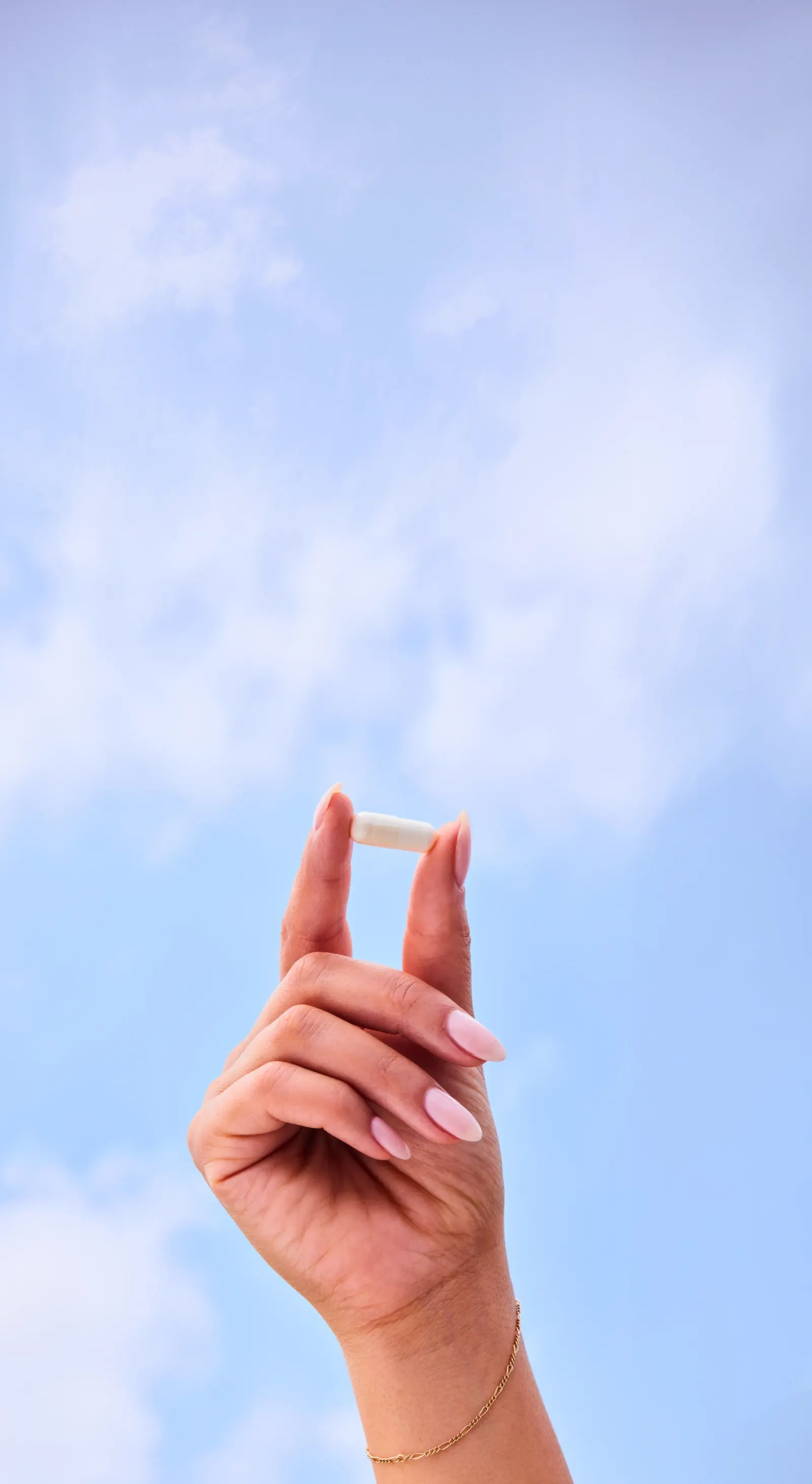 Hand holding a single supplement capsule against a bright blue sky