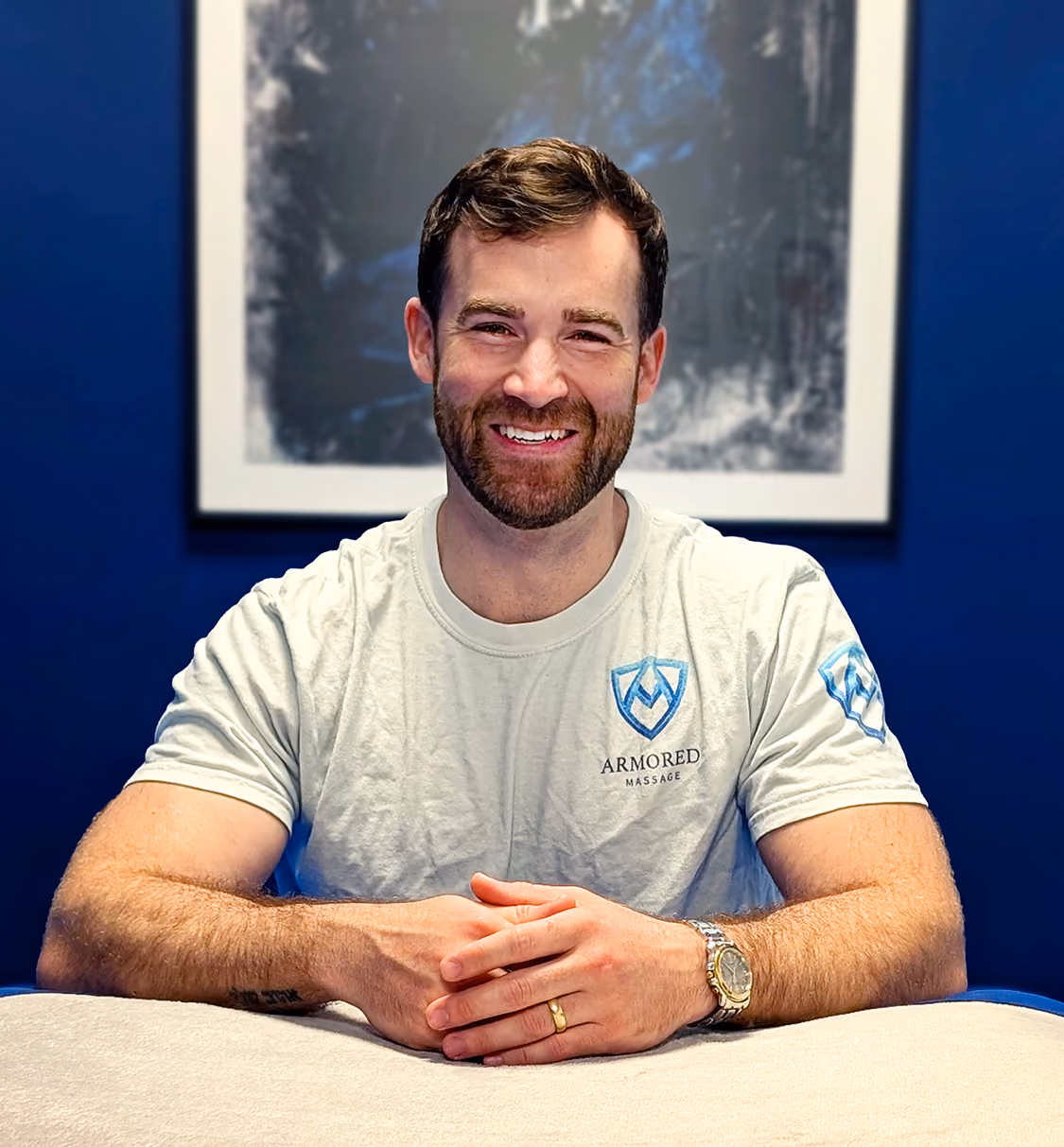 Stephen Preyer smiling at the camera sitting behind his massage table, he looks friendly and cheerful, his hands are resting on the massage table, and behind him is a wall with an image of a knight behind him with a cape.
